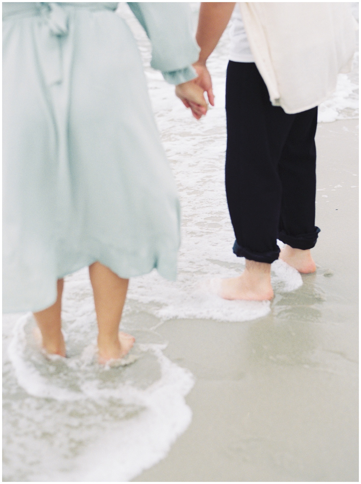 Couple during their engagement session holding hands and feet in the sand and ocean